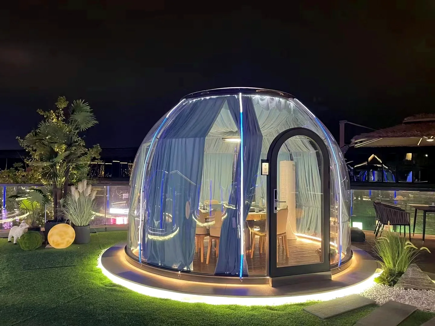 Round 3.5 m High clear polycarbonate dome lit at night on a roof garden, curtains drawn and a dining table inside